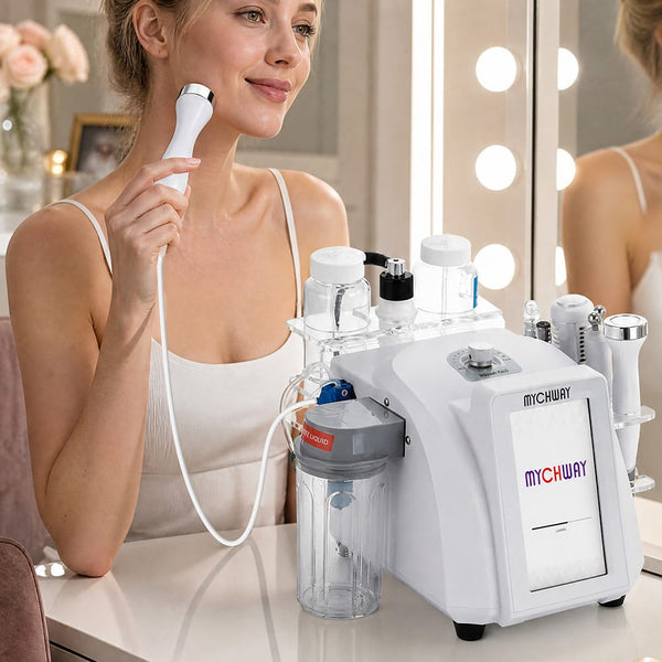 A woman in a white tank top uses a white MYCHUAY facial treatment device with a handheld wand attachment on her face, set in a bright vanity room with a mirror and light bulbs in the background.