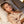 A woman with closed eyes rests on a bed with white pillows and sheets, wearing a satin pajama shirt in a soft champagne beige color.