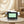 A modern facial treatment device labeled FACEVO sits on a round table in a softly lit spa-like room. The device has a white and black body with a green welcome screen and two white handheld applicator heads attached to the sides.