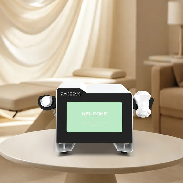 A modern facial treatment device labeled FACEVO sits on a round table in a softly lit spa-like room. The device has a white and black body with a green welcome screen and two white handheld applicator heads attached to the sides.