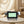 FACEEVO facial cleansing device with two white and black brush heads, displayed on a round beige table in a softly lit spa-like room.