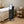 A black mobile skincare or beauty treatment machine with a touchscreen display and caster wheels, placed beside a neatly made spa bed in a treatment room. A wooden cart with towels and bottles stands nearby.